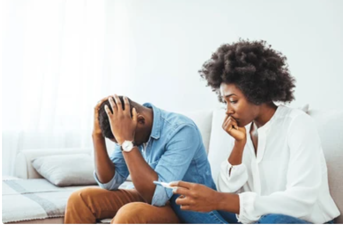 Concerned couple sitting on couch discussing fertility challenges - woman looking at pregnancy test while man supports her with head in hands showing emotional stress of fertility journey