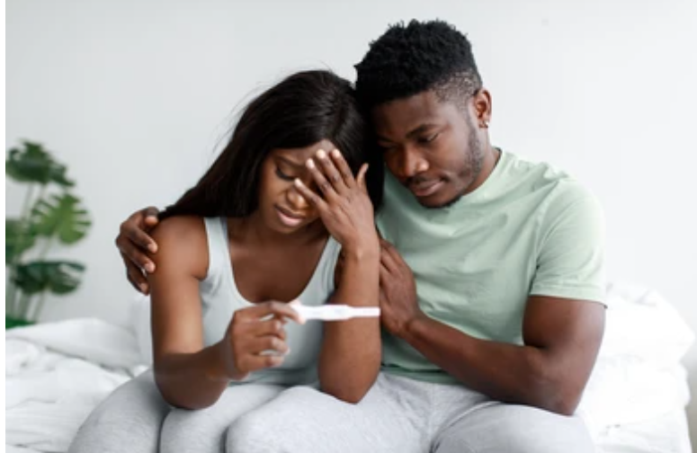 Couple on bed dealing with fertility disappointment - woman covering face in distress while holding pregnancy test, man providing emotional support and comfort during difficult fertility journey